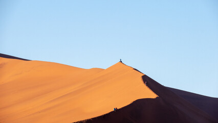 Fototapeta na wymiar Tourists walking and sitting on the edge of a red sand dune at Sossusvlei National Park, a popular tourist destination in Namibia