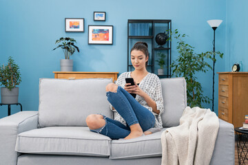Contented woman sits on the sofa in the living room holding her phone in her hands, a girl sends...