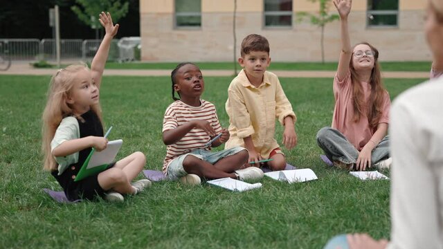 Diverse school children raising hands during lesson outdoors