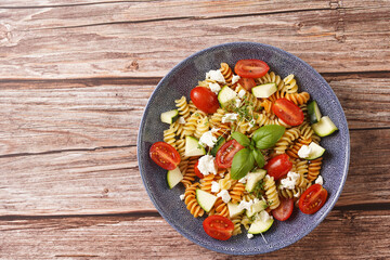 Italian pasta fusilli tricolore with cherry tomatoes, zucchini and feta cheese in a blue shallow bowl on a red napkin with black silverware on a wooden surface