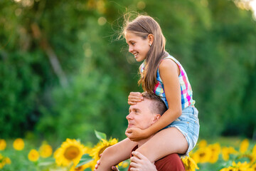 A young father plays with his daughter in a field of sunflowers. A little girl sits on her father's neck and laughs. Family summer vacation outside