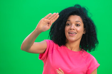 Portrait of young female African American is waving hand hello and smiling. Black woman with curly hair in pink tshirt poses on green screen in the studio. Close up.