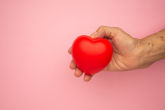 Hand of a senior woman holding a red heart shape with a pink background. Close-up photo. Aged people and healthcare concept