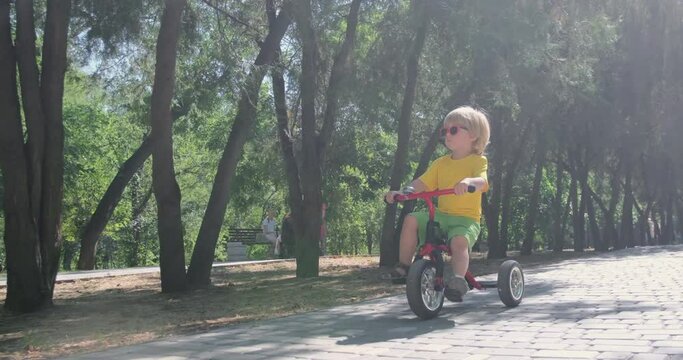 Adorable small blond boy with sunglasses in stylish clothes rides modern tricycle along paved road in green park on sunny day