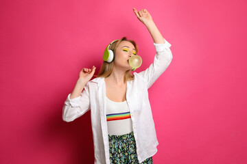 Fashionable young woman with bright makeup and headphones blowing bubblegum on pink background