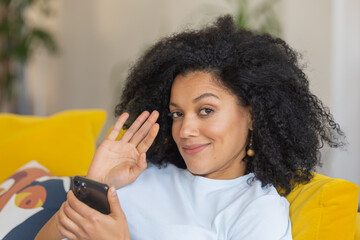 Fototapeta premium Portrait of a young African American woman looking at the camera and waving hand hello. Brunette with curly hair sitting on yellow sofa in a bright home room. Close up.
