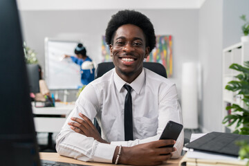 Cheerful man dressed in white shirt tie is sitting at desk in front of computer holding phone in hand waiting for call boss manager is smiling at camera in background other people working in office