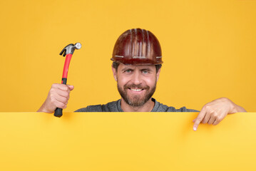 happy mature man handyman in helmet behind yellow paper for copy space hold hammer, presenting