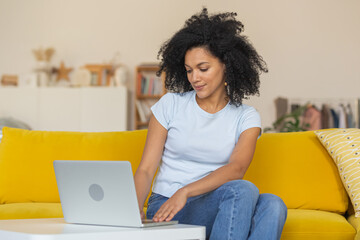 Obraz na płótnie Canvas Portrait of a young African American woman typing on laptop keyboard. Brunette with curly hair sitting on yellow sofa in a bright home room. Close up.