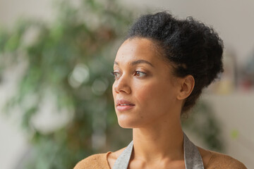 Portrait of a young African American woman looking to the side. Young female owner of an online store in an apron posing against on a blurred background. Close up.