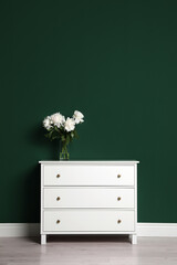 Modern white chest of drawers with beautiful bouquet near green wall indoors