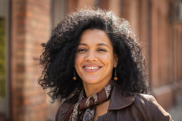 Portrait of stylish young African American woman smiling and looking at camera. Brunette with curly hair in brown leather jacket posing on street against backdrop of blurred brick building. Close up.