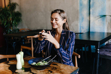 Happy girl taking photo of her salad. Indoor portrait of smiling brunette woman having fun during dinner.