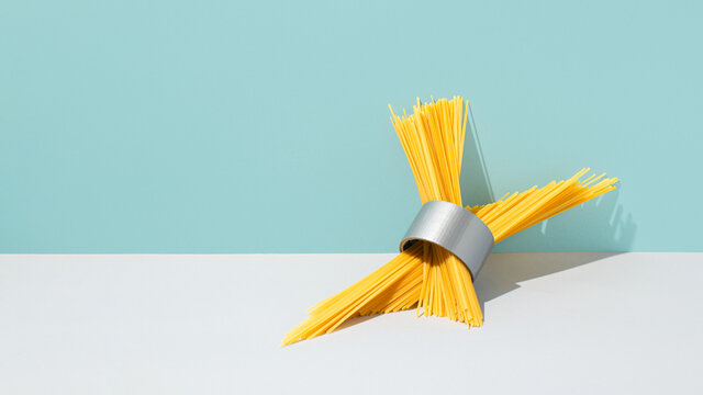 Raw spaghetti and duct tape roll composition against blue background. Abstract yellow pasta with grey adhesive tape creative concept. Surreal italian food art.