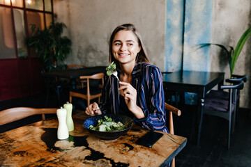 Smiling dark-eyed girl eating salad in cafe. Cheerful female model enjoying vegetarian dish.