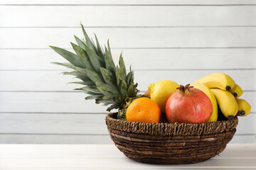 Wicker bowl with different ripe fruits on white wooden table. Space for text