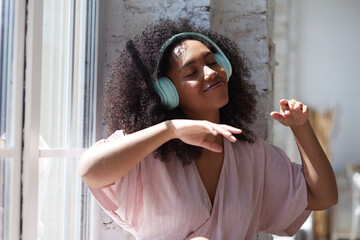 Happy hilarious black multiracial lady wearing headphones near the window listening to modern hip hop singing song with eyes closed resting in comfortable apartment at home