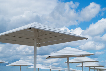 Wooden canopies in form of quadrangular pyramidal umbrellas, painted white, installed on beach to create shadow for vacationers, leaving rows in perspective against background of cloudy blue sky.