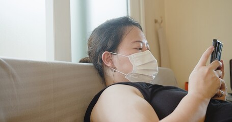 Attractive Asian woman wearing protective medical face mask using smartphone relaxing on sofa at home, Health care and medical covid-19 concept
