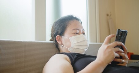 Attractive Asian woman wearing protective medical face mask using smartphone relaxing on sofa at home, Health care and medical covid-19 concept