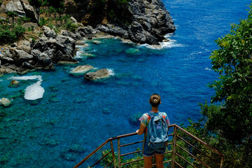 Young woman wearing tie dye t-shirt and a backpack enjoying the view of blue lagoon, clear turquoise water. Female traveler standing on a cliff over mediterranean sea coastline. Copy space, background
