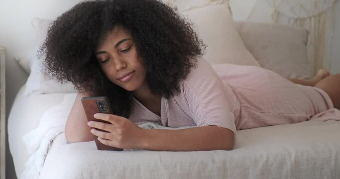 A relaxed, quiet black woman lies on her bed and enjoys social networking on her smartphone. An uplifted mixed-race girl enjoys browsing the news and messages on her phone.