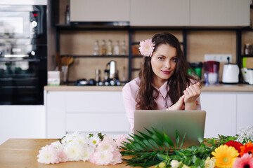 Young brunette woman with long wavy hair working on laptop with flowers on the desk with kitchen in background. Talented florist developing online sales getting ready for workshop.