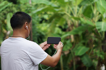  Side view of Asian man holds blank screen smartphone for text and listen to music. Concept : Wireless technology smart device internet using in daily life. Nature background.         