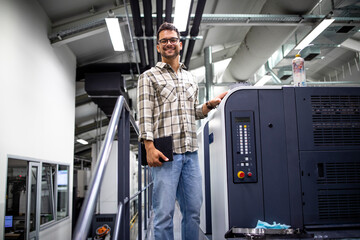 Portrait of male designer standing by printing machine and controlling imprint quality in printing...