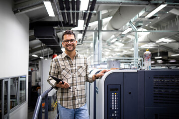 Portrait of print manager standing by large offset printing machine in print factory. - obrazy, fototapety, plakaty