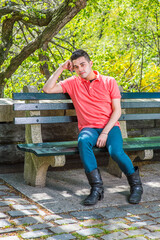 Fototapeta premium Dressing in a red Polo shirt, green pants and long leather boots, one arm supporting his head, a young handsome guy is sitting on a long chair in the morning of a spring season, relaxing.