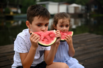 Handsome boy eating slice of delicious sweet juicy watermelon, sitting next to his younger sister. Enjoying summer day on the countryside pier.