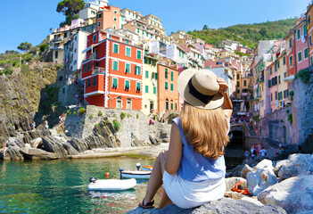 Beautiful young woman sitting on rock enjoying fantastic view of Riomaggiore colorful village in...