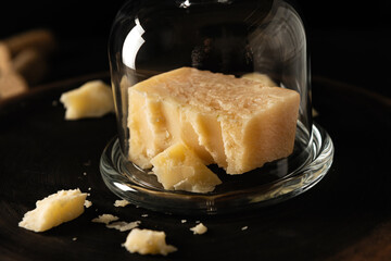 sliced parmesan cheese with chipped pieces, covered with a glass jar, on a dark background