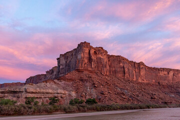 The brilliant colors of a sunrise reflecting on clouds over the top of a canyon wall with a river in the foreground in Utah.