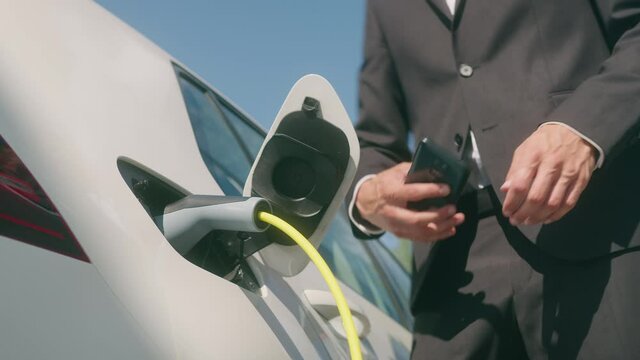 Handheld shot of a Caucasian man in a black suit, standing near an electric car that is charging and making time adjustments on a smartphone