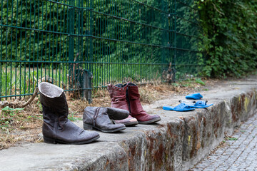 Alte Stiefel und Sandalen stehen aufgereiht auf einer niedrigen Mauer mit einem grünen Metallzaun im Hintergrund.