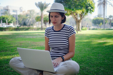 Asian woman is sitting on the grass in the park, working on her laptop. video communication, work, study and e-commerce concept