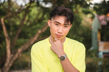 Portrait of a handsome asian guy in a yellow shirt and pearl necklace posing at a park.