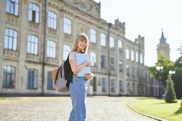 Adorable happy young blonde university or high school girl student with backpack and laptop wearing...