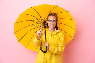 Teenager Russian girl with rainproof coat and umbrella isolated on pink background showing and...