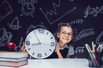Pupil girl with big clock at the black chalkboard in classroom.