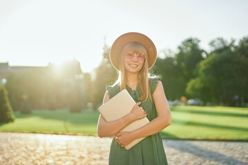 Adorable young blonde university or college student girl with laptop wearing green dress and hat in university campus. Happy female school or student, educational concept. High quality image