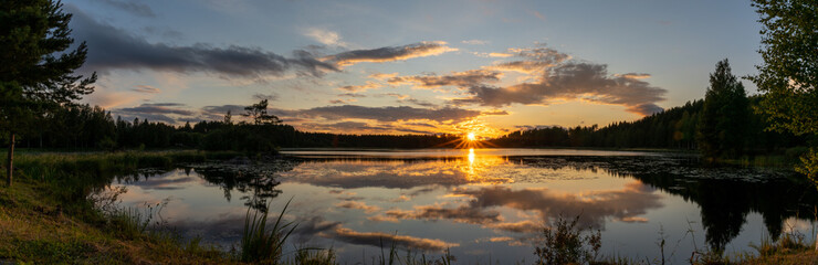 panorama of a colorful sunset reflected in a calm lake landscape with green forest and reeds
