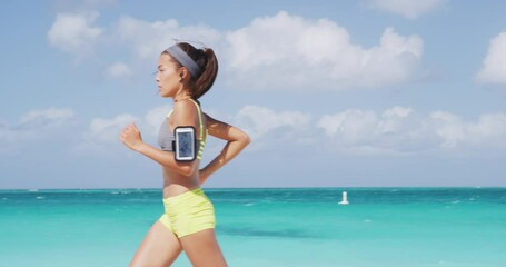 Running athlete woman jogging on beach listening to podcast with wireless earphones and mobile phone. Asian runner doing outdoor exercise healthy active lifestyle