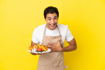 Restaurant waiter holding waffles over isolated yellow background smiling a lot