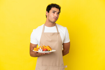 Restaurant waiter holding waffles over isolated yellow background and looking up