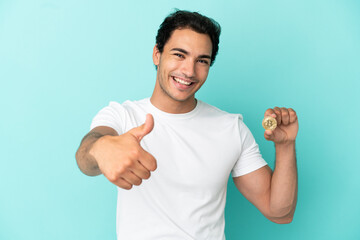 Young man holding a Bitcoin over isolated blue background with thumbs up because something good has happened