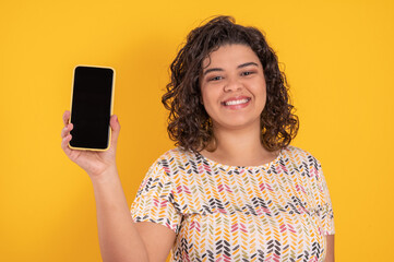 brazilian young woman holding and showing a cellphone to the camera on a yellow background