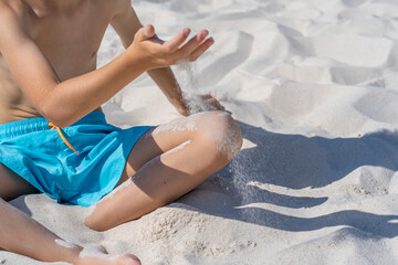 Hands of a young boy in blue shorts sits on the white sand on the beach. Plays with white sand. High quality photo
